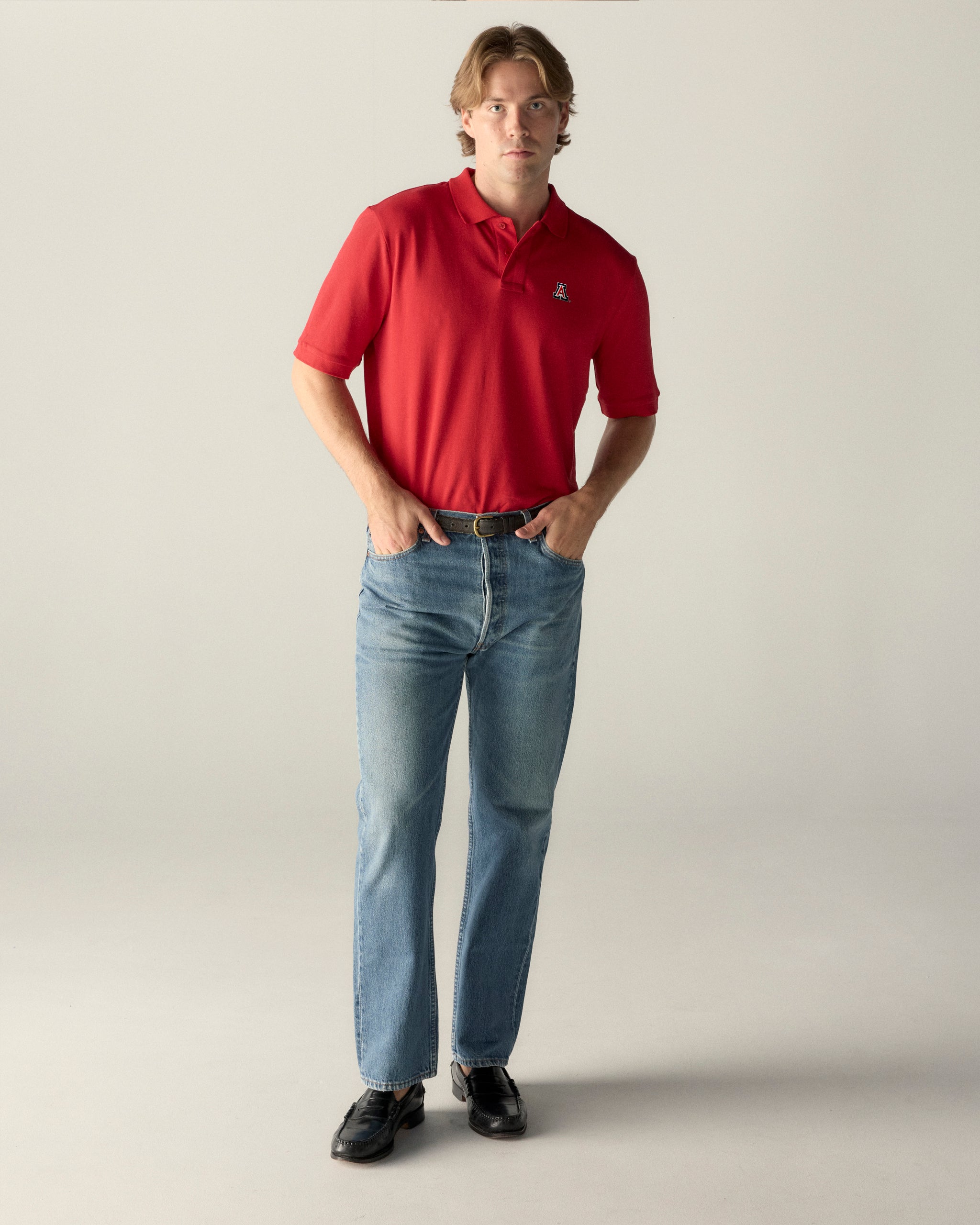 Man wearing red collegiate polo shirt with embroidered logo and blue jeans, standing indoors. Class Trip | Officially Licensed Collegiate Apparel