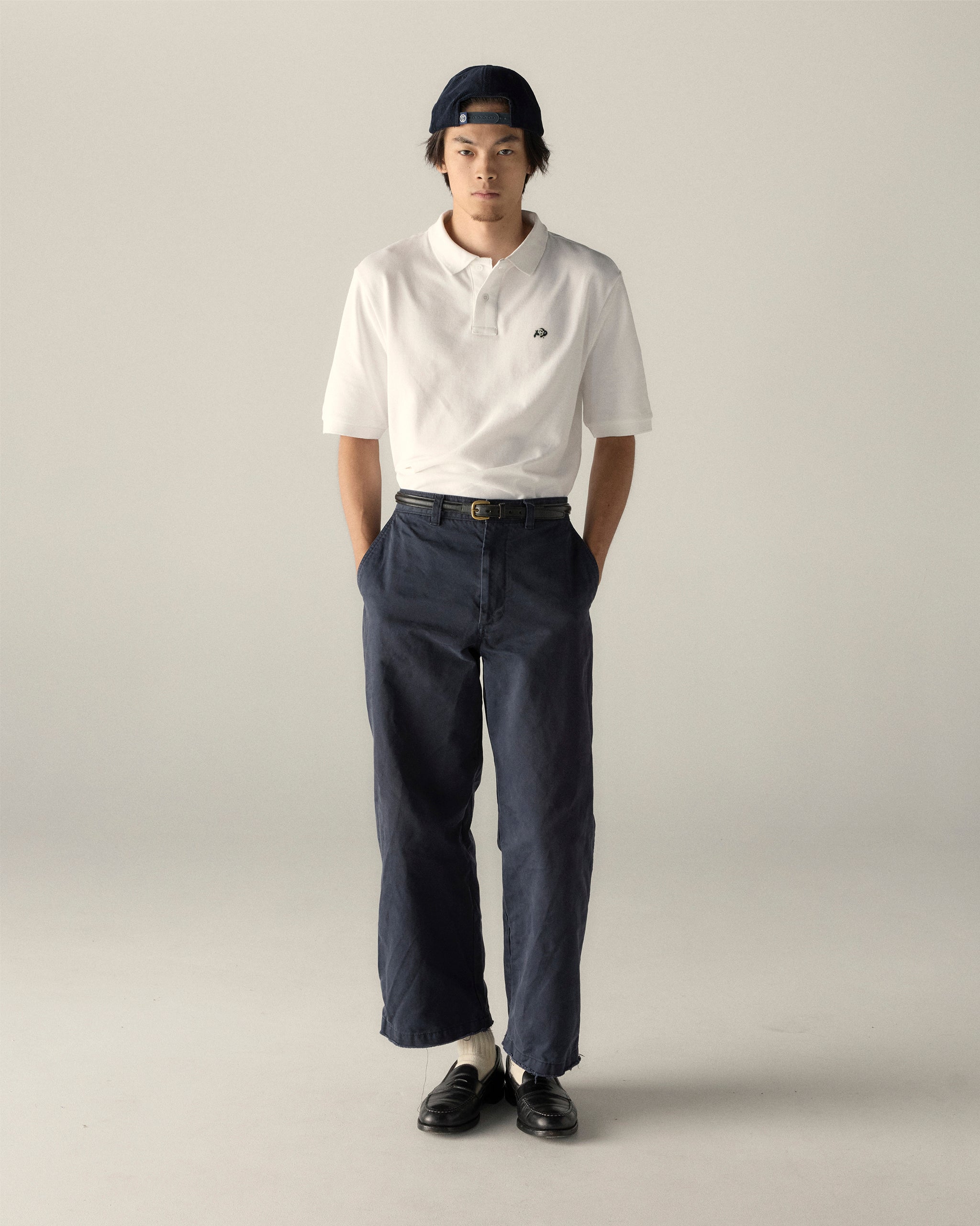 Man wearing a white pique polo shirt, navy pants, black loafers, and a backwards cap, standing against a plain background. Class Trip | Officially Licensed Collegiate Apparel