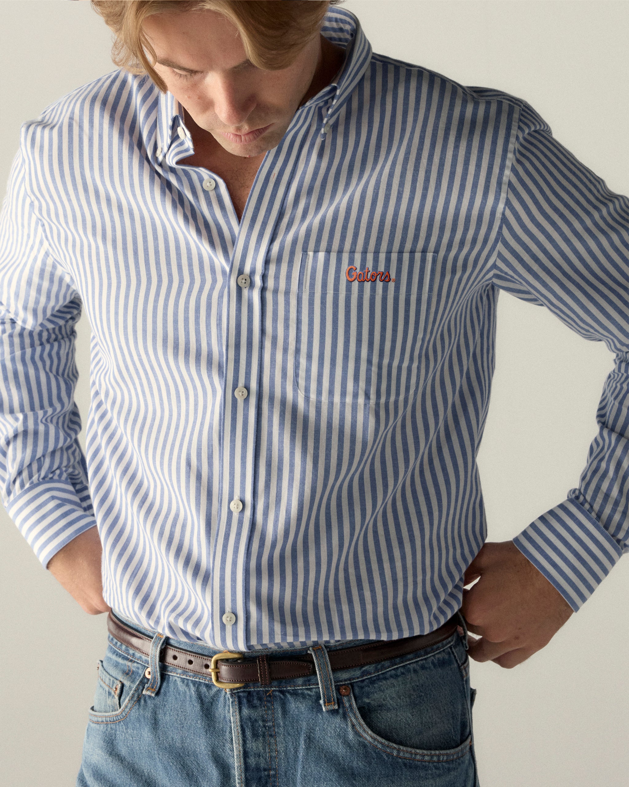 Man wearing blue and white striped poplin button-down shirt with embroidered collegiate logo, casual jeans. Class Trip | Officially Licensed Collegiate Apparel