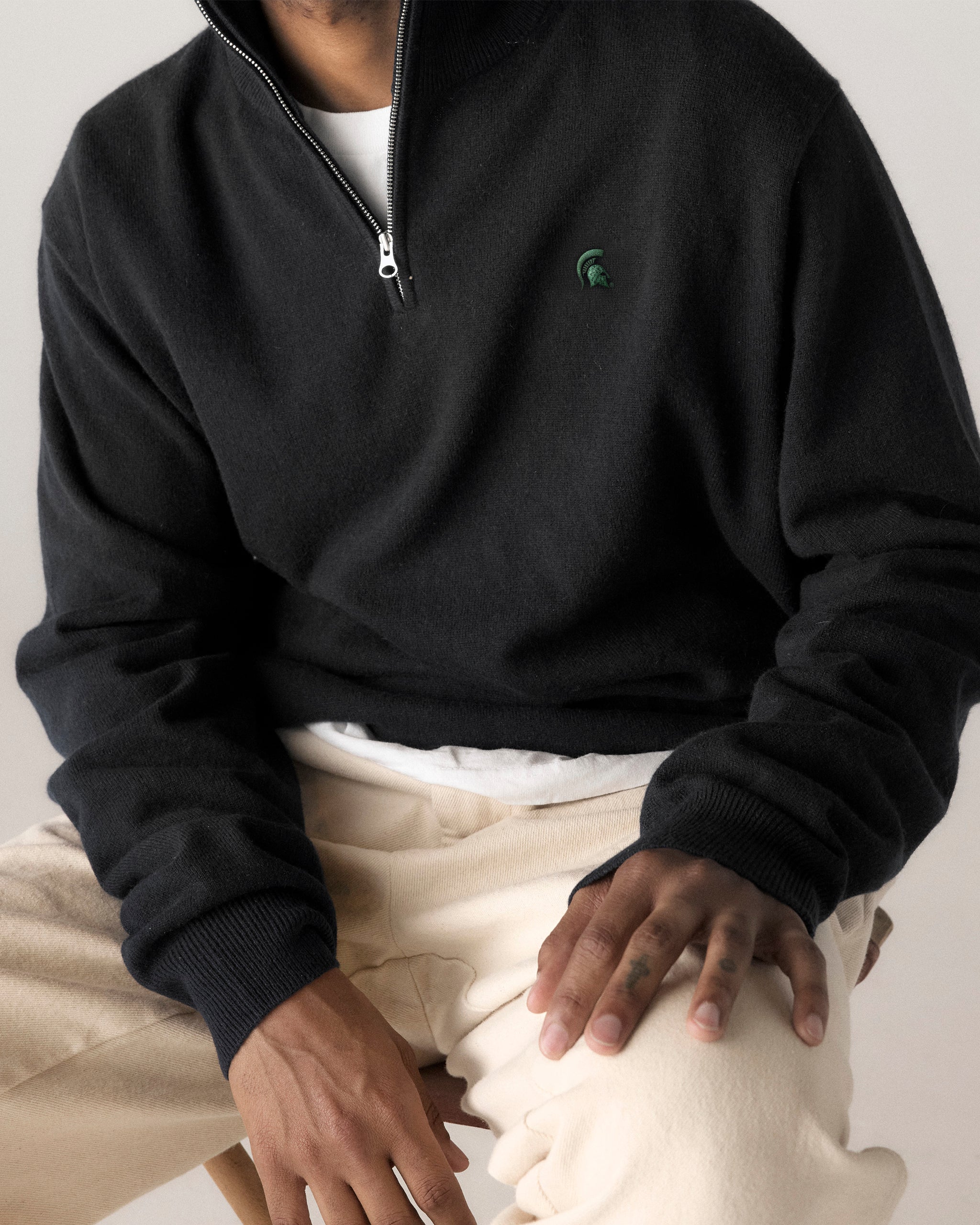 Man wearing black quarter-zip pullover with green collegiate logo, beige pants. Class Trip | Officially Licensed Collegiate Apparel