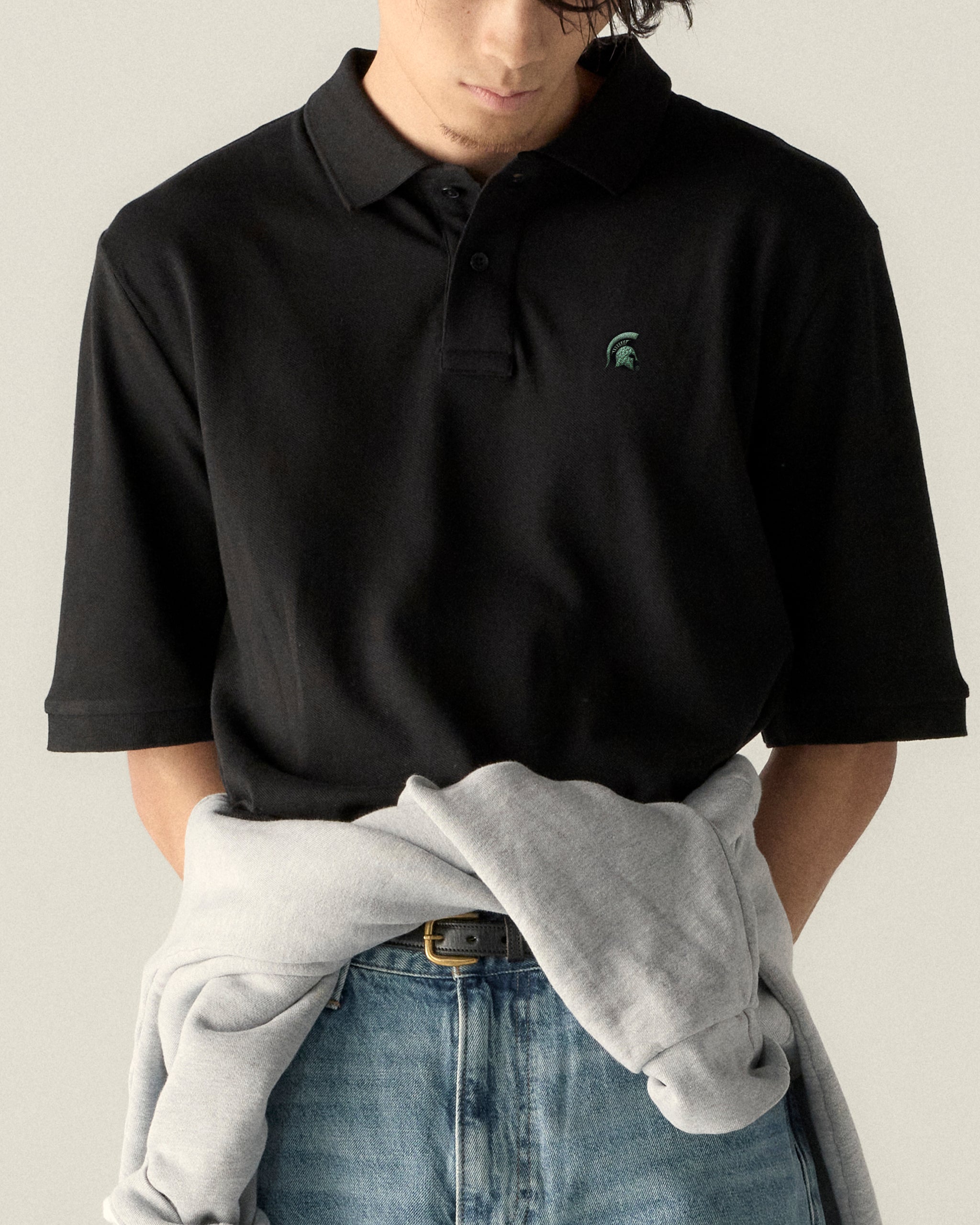 Black collegiate polo shirt with green Spartan logo, paired with jeans and a gray sweatshirt. Class Trip | Officially Licensed Collegiate Apparel