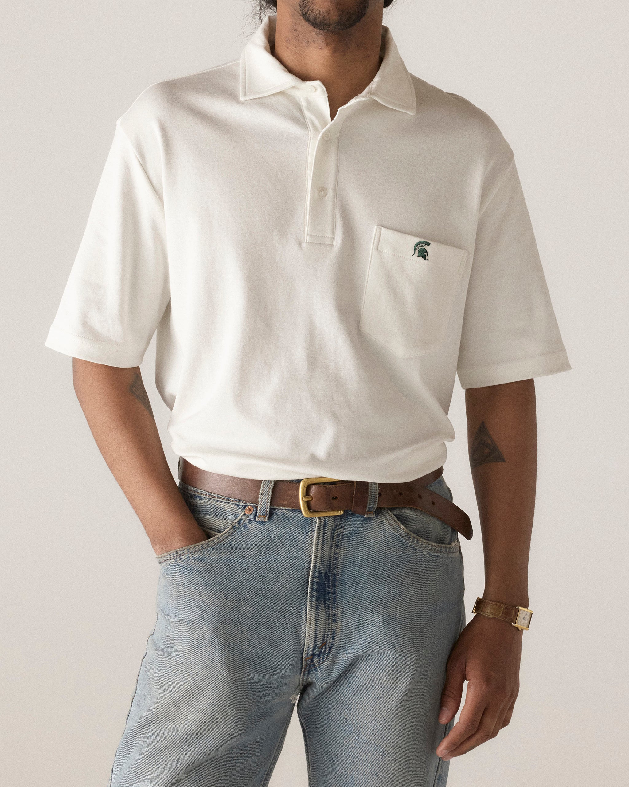 White collegiate polo shirt with pocket logo, styled with jeans. Class Trip | Officially Licensed Collegiate Apparel