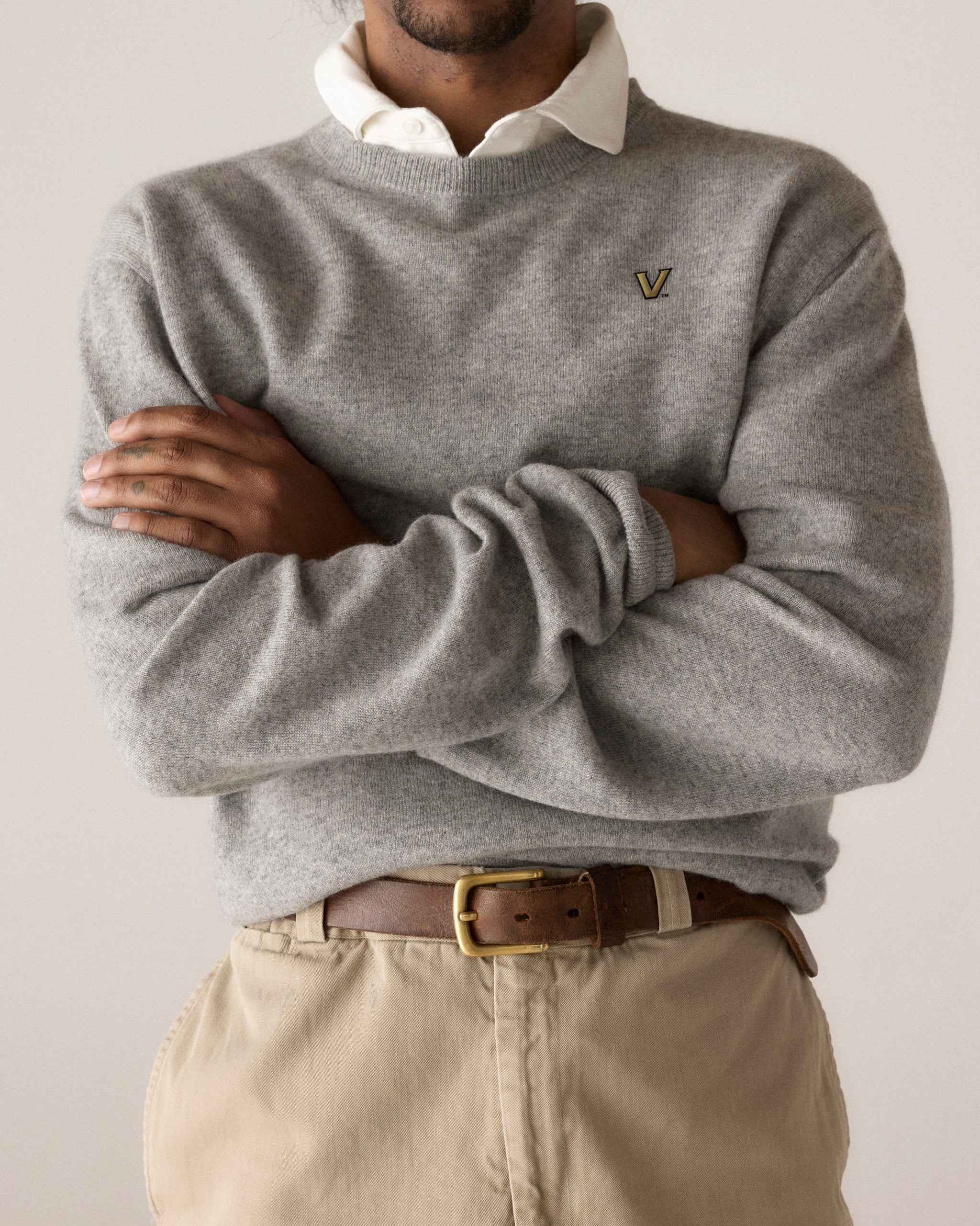 Man wearing a grey merino crewneck sweater with collegiate V logo, beige pants, and a brown belt. Class Trip | Officially Licensed Collegiate Apparel