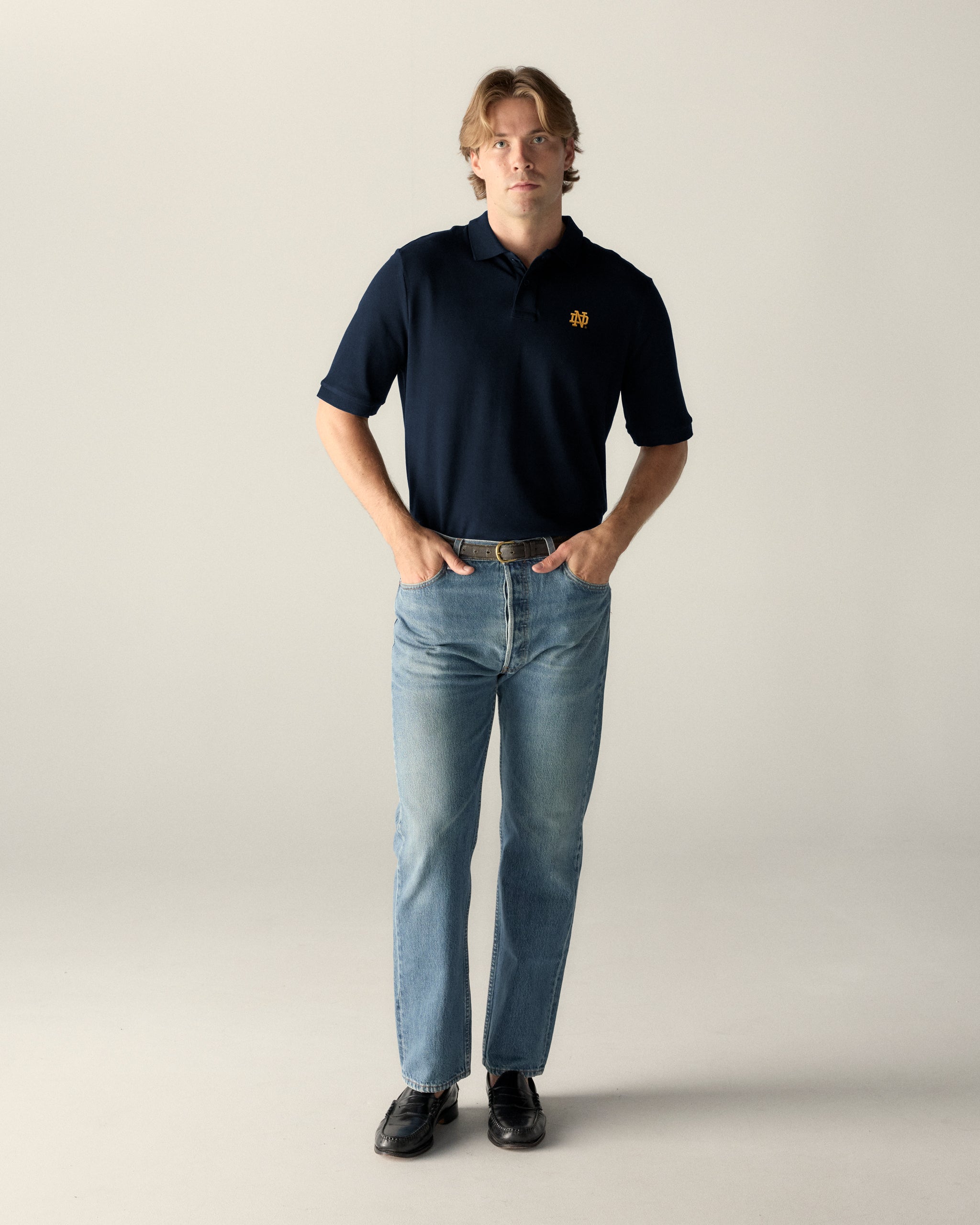 Man wearing navy collegiate polo with gold embroidered crest and blue jeans, Class Trip | Officially Licensed Collegiate Apparel