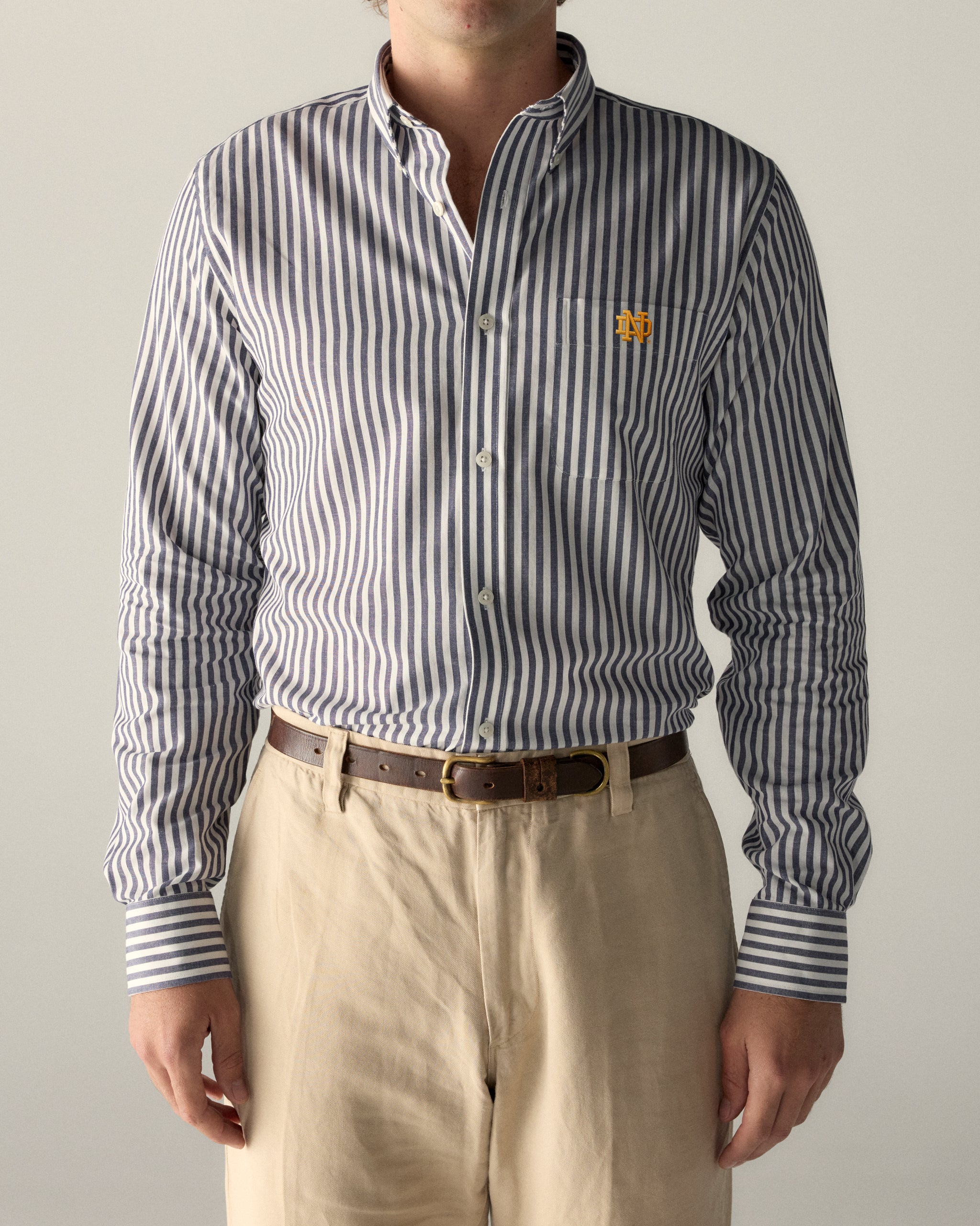Men's striped button-down shirt with embroidered collegiate logo, tucked into khaki pants. Class Trip | Officially Licensed Collegiate Apparel