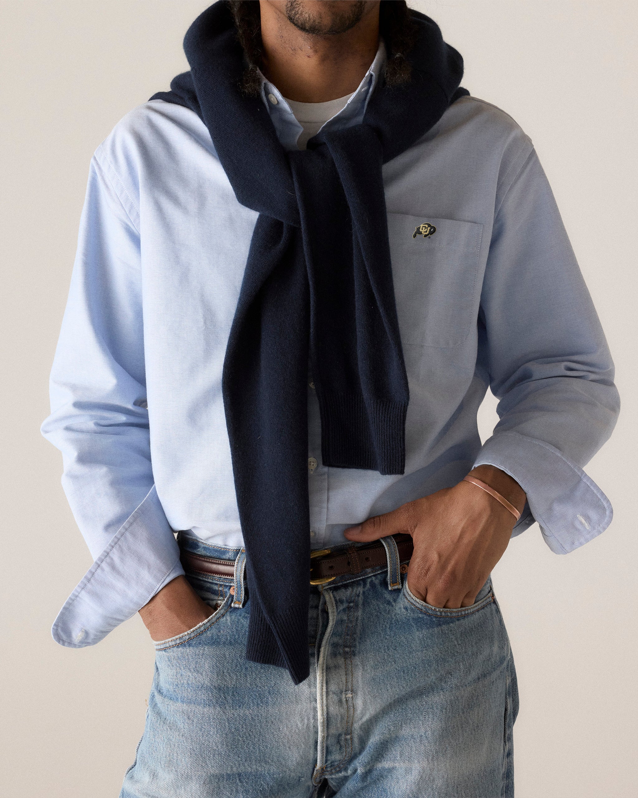 Man wearing light blue oxford shirt with collegiate logo, navy sweater, and jeans. Class Trip | Officially Licensed Collegiate Apparel