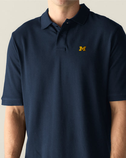 Navy blue pique polo shirt with yellow collegiate M logo on chest, short sleeves. Class Trip | Officially Licensed Collegiate Apparel
