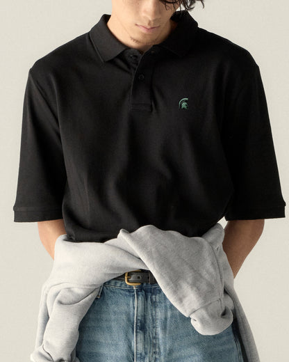 Black collegiate polo shirt with green Spartan logo, paired with jeans and a gray sweatshirt. Class Trip | Officially Licensed Collegiate Apparel