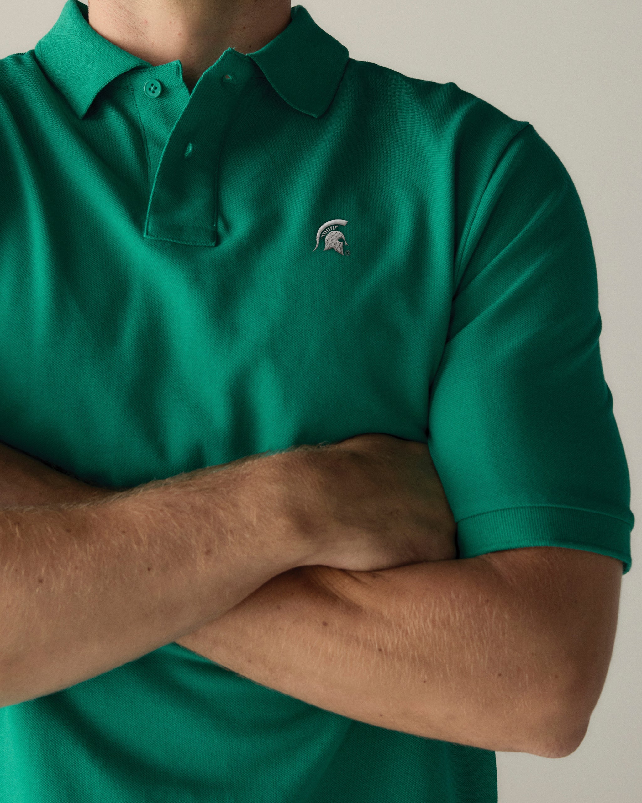 Green collegiate pique polo shirt with university logo, arms crossed, front view. Class Trip | Officially Licensed Collegiate Apparel