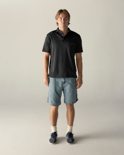 Man modeling black collegiate polo shirt with pocket and denim shorts, casual style. Class Trip | Officially Licensed Collegiate Apparel