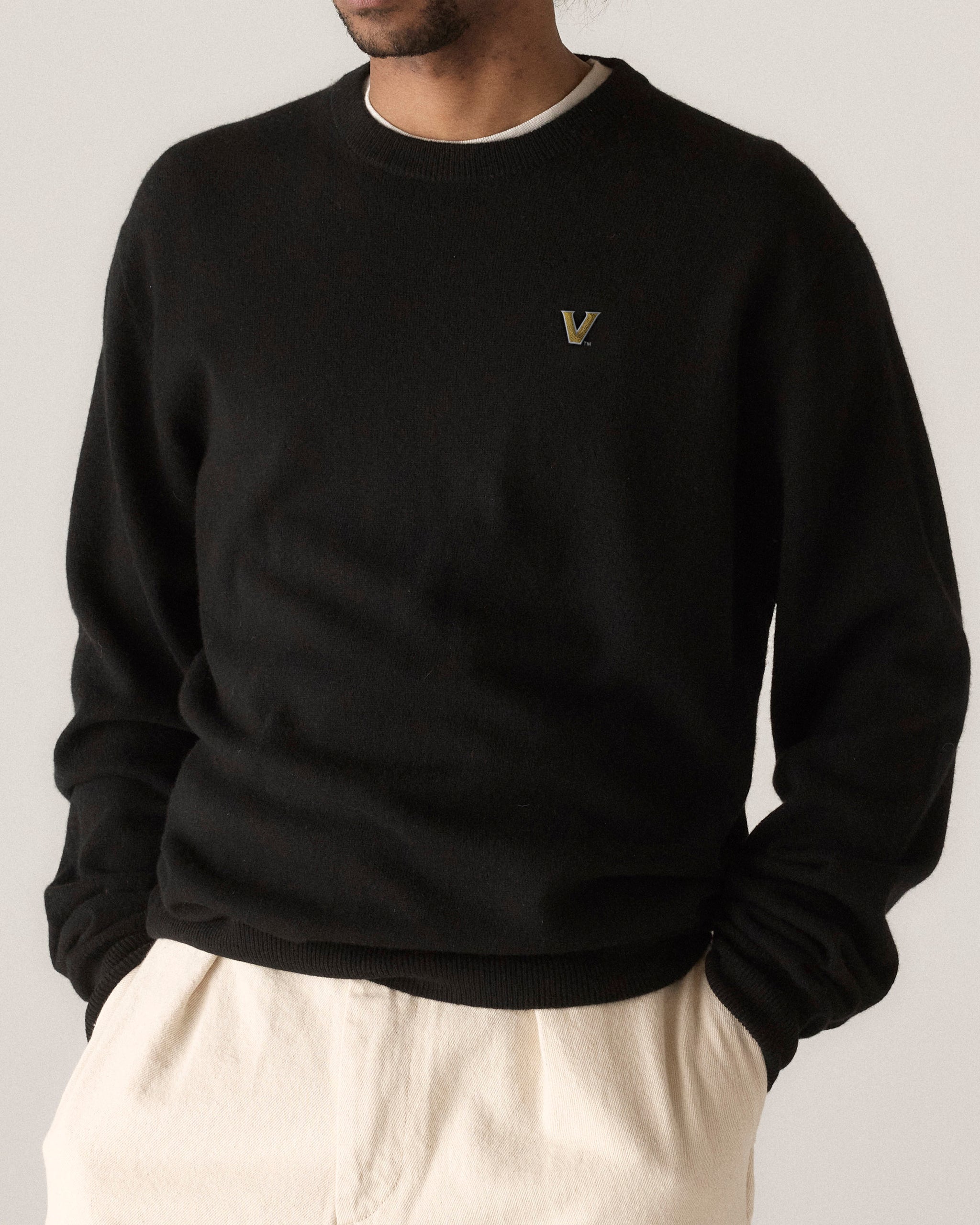 Man wearing black merino sweater with gold collegiate V logo, cream pants. Class Trip | Officially Licensed Collegiate Apparel